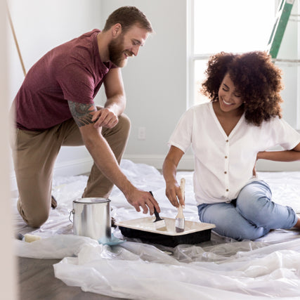 Paint & SuppliesA couple kneels on a plastic-covered floor, smiling as they paint a room. A paint can and tray are beside them, with a ladder in the background.