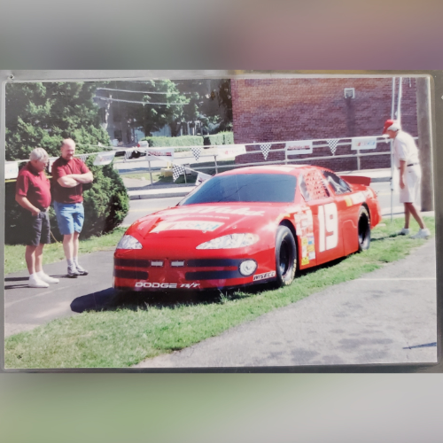 A red race car with the number 19 is parked on a lawn beside a building. Three men in casual clothing stand nearby, observing the car.