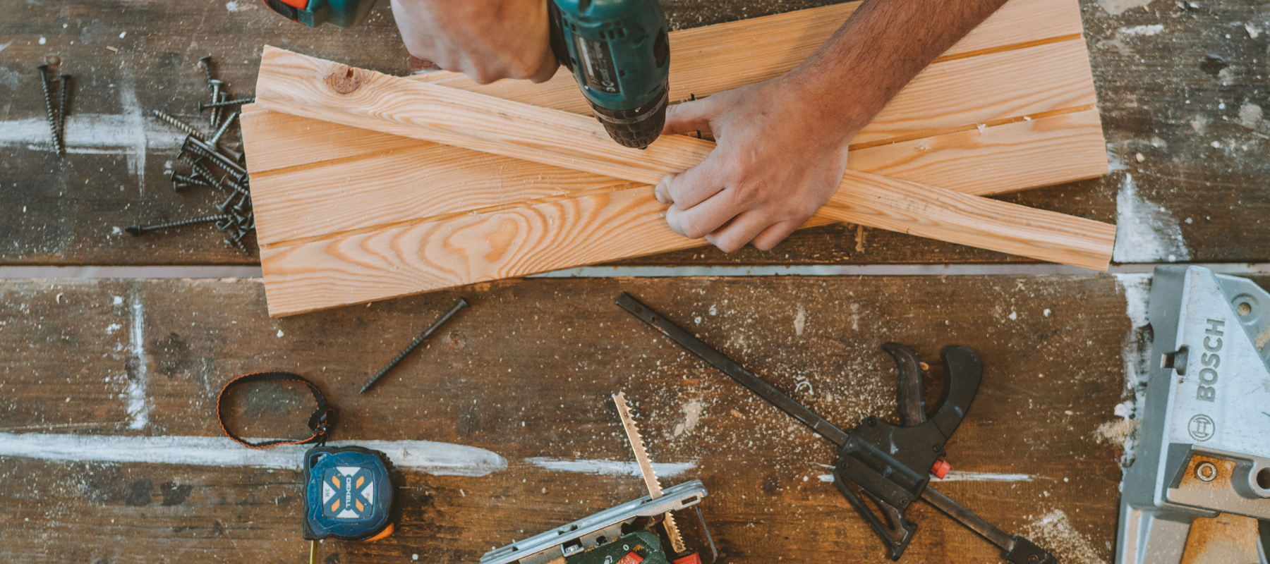 Hands using a power drill on two pieces of wooden planks on a workbench. Nearby are tools like a tape measure, clamp, and various screws.
